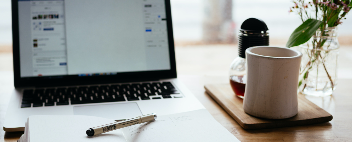 Ein Bild von einem Laptop mit geöffneter Webseite. Davor liegt ein offenes Notizbuch mit Stift und eine Kaffeetasse.